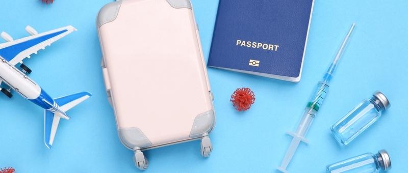 Model aeroplane, suitcase, syringes, passport, and vaccine bottles against blue background.