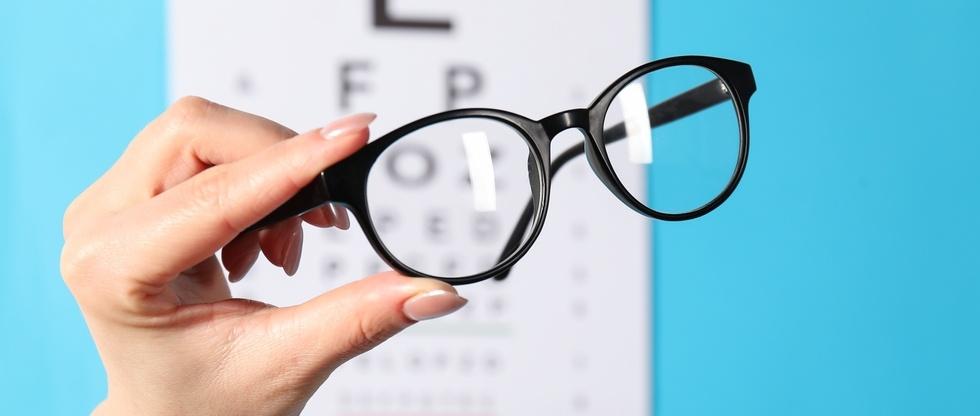 Woman holding glasses against vision test chart on light blue background, closeup