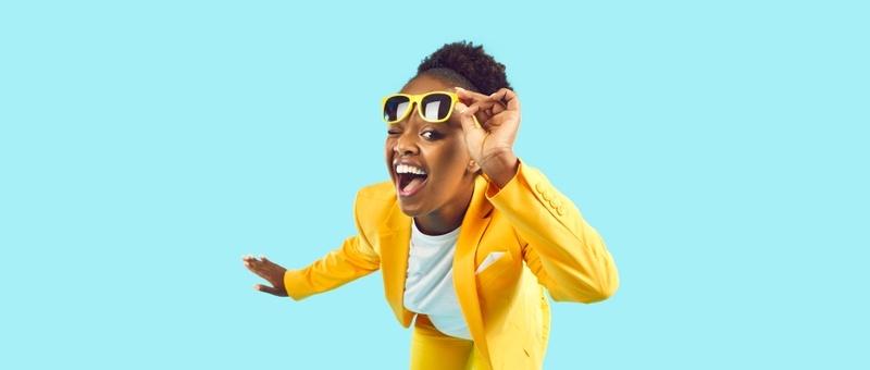 Woman wearing white top and yellow blazer winks beneath a pair of yellow sunglasses against a bright blue background.