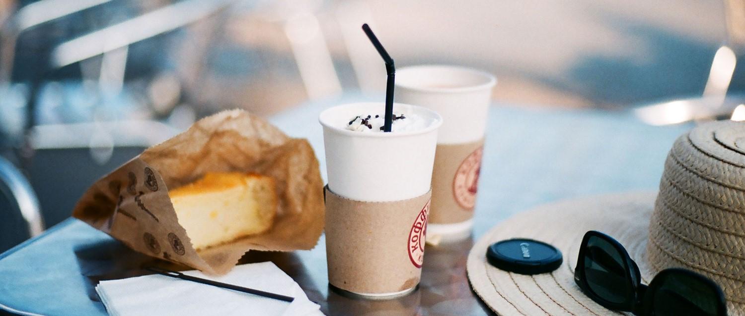 Coffee cups with whipped cream, pastry in paper bag, sunglasses and straw hat on café table.