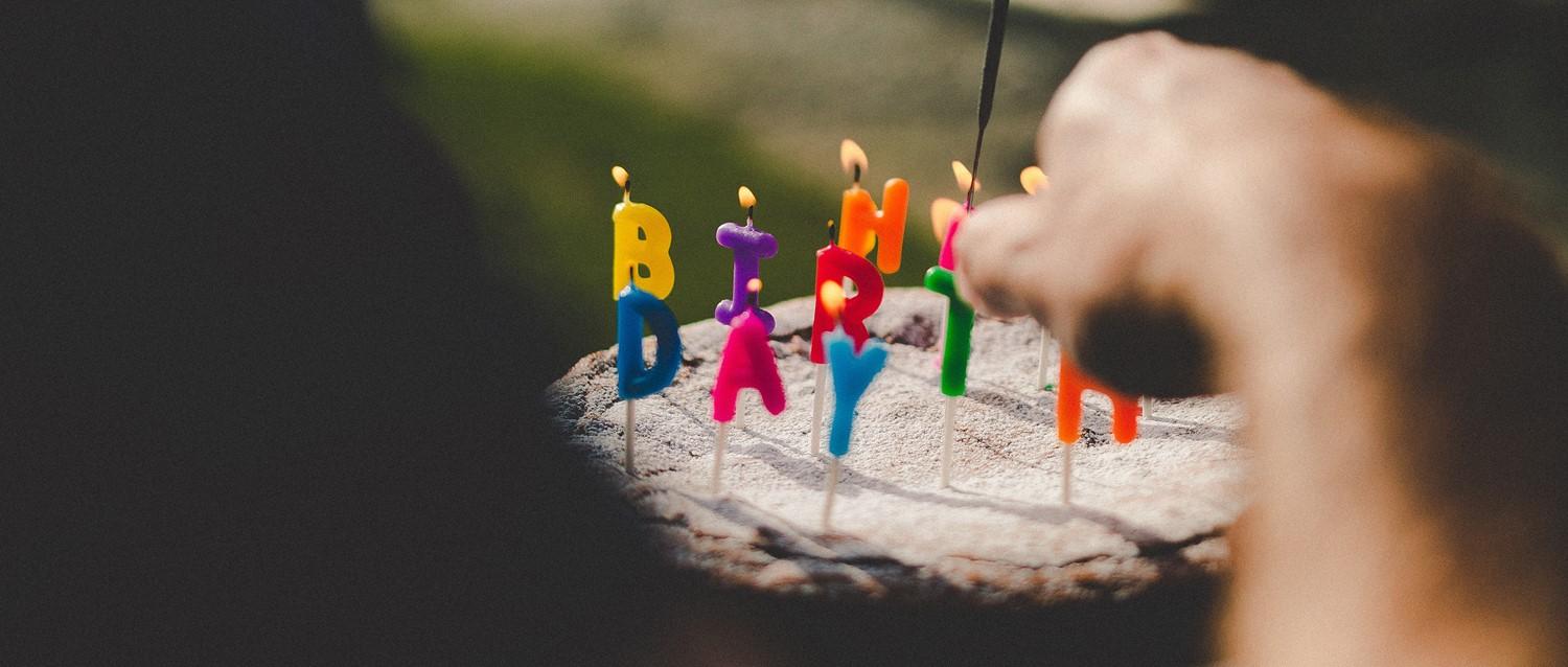 Colourful "BIRTHDAY" letter candles with lit flames on a cake, with a hand visible lighting one of the candles.
